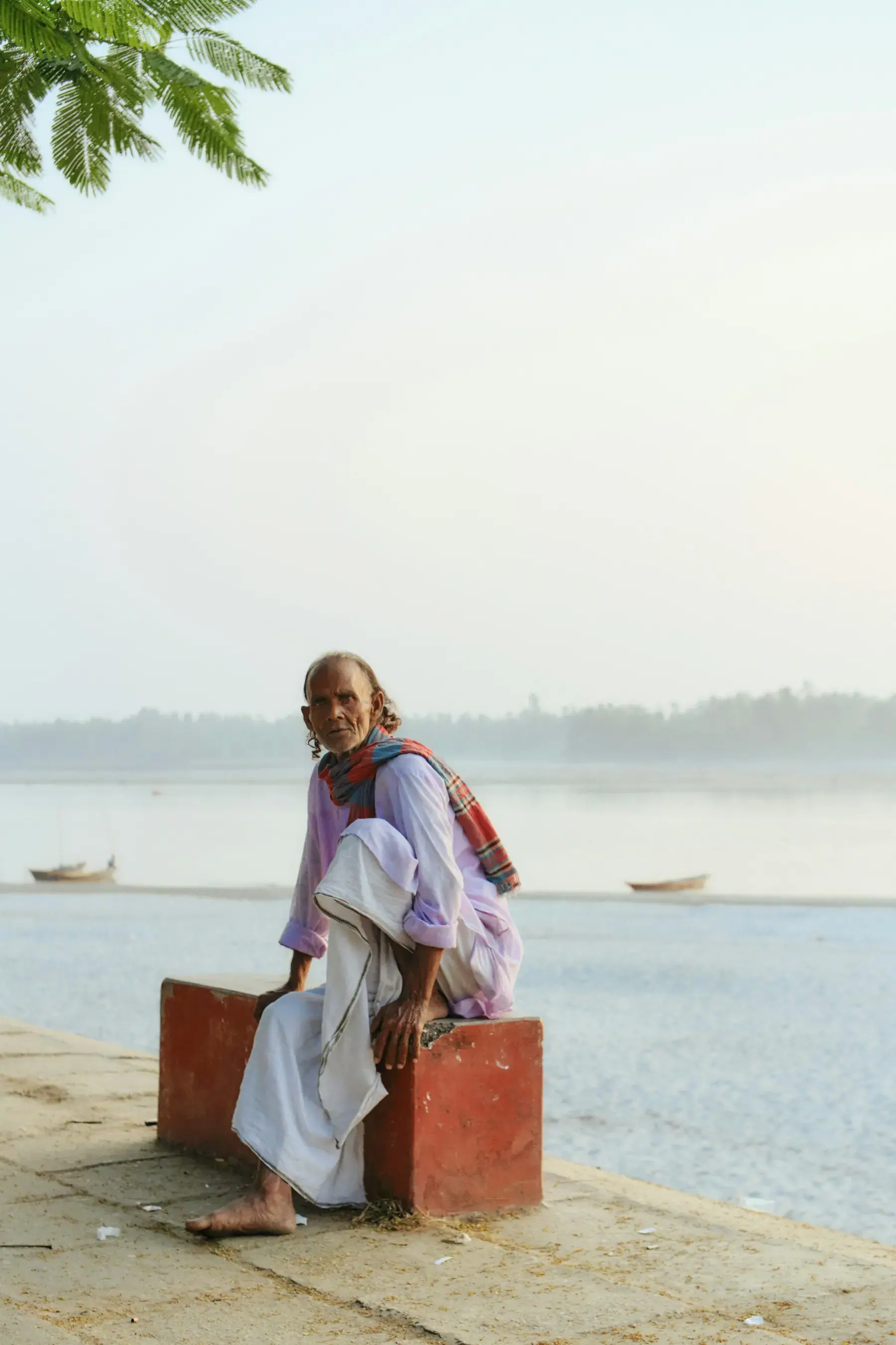 An elder seated beside a Bangladesh river at sunrise